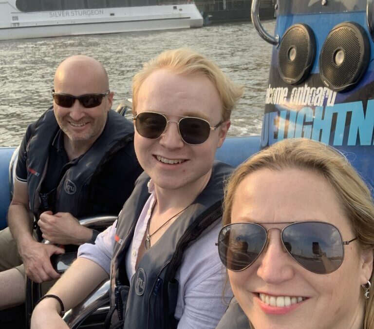 Three individuals wearing sunglasses and life jackets on a boat, with a vessel in the background.