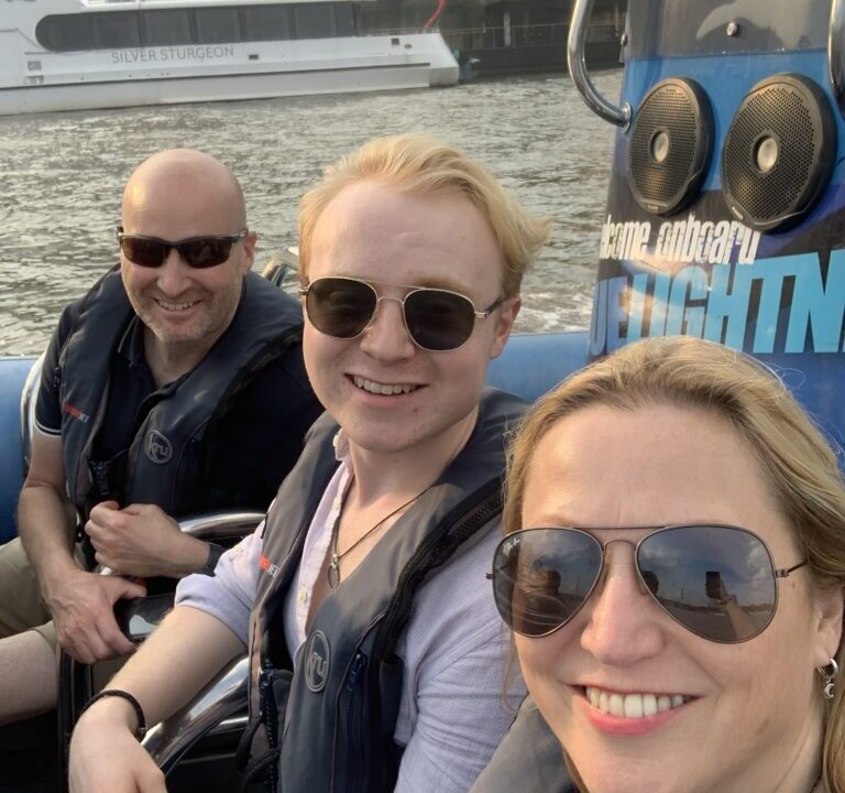 Three individuals wearing sunglasses and life jackets on a boat, with a vessel in the background.