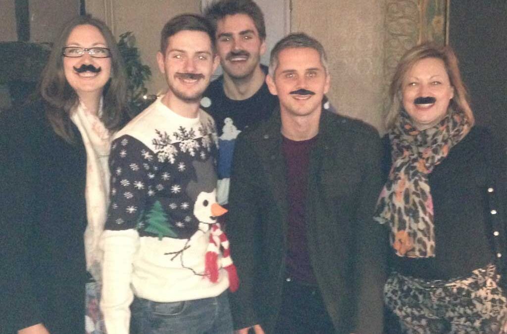 Five individuals wearing fake mustaches and festive attire posing together for a photo.