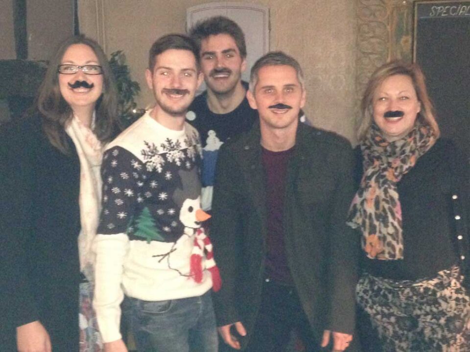 Five individuals wearing fake mustaches and festive attire posing together for a photo.