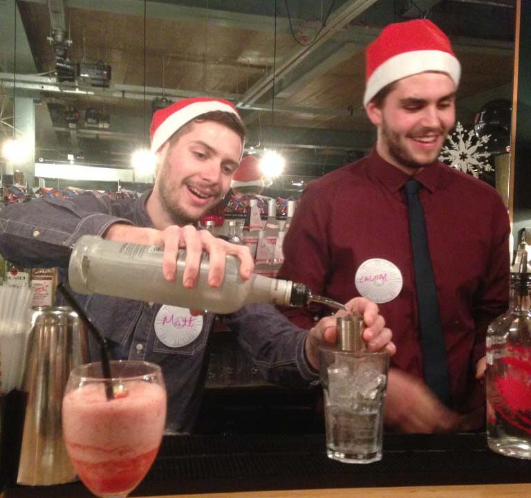 Two bartenders wearing Santa hats mixing drinks at a bar counter with cocktails in the foreground.