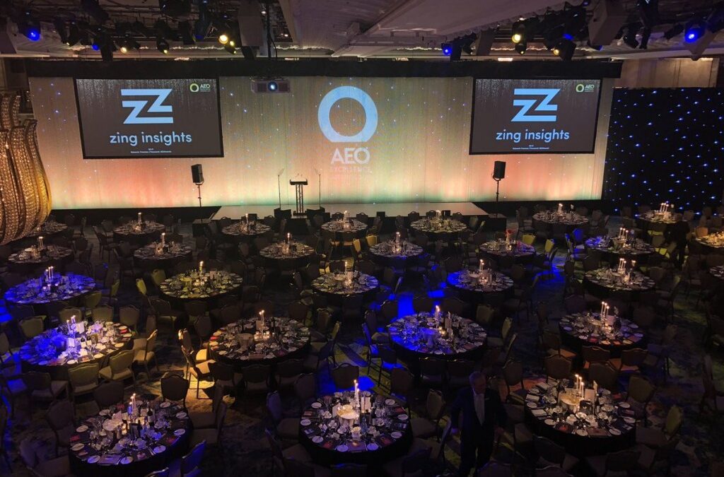 A large event space with round tables set for dining and projector screens displaying logos.