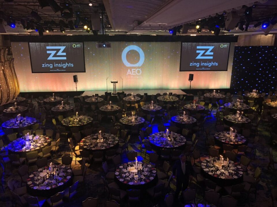 A large event space with round tables set for dining and projector screens displaying logos.